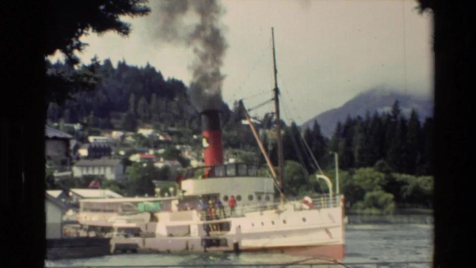 1982 New Zealand 8mm frame showing the Earnslaw steamer near shore, with black smoke, lake water, hills, buildings, and passengers on deck.