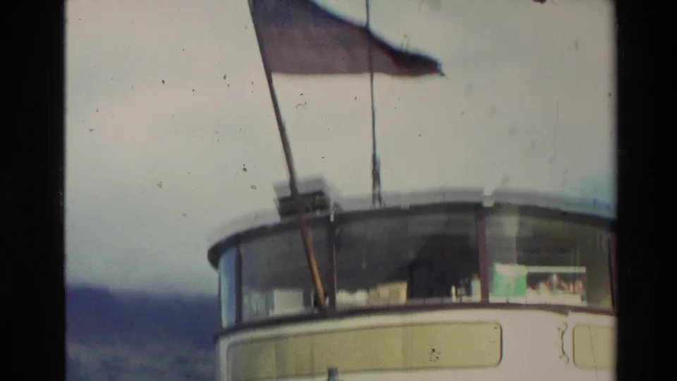1982 New Zealand 8mm frame looking up at the steamer upper deck, with a flag, mast, cabin windows, and soft lake and hill background.