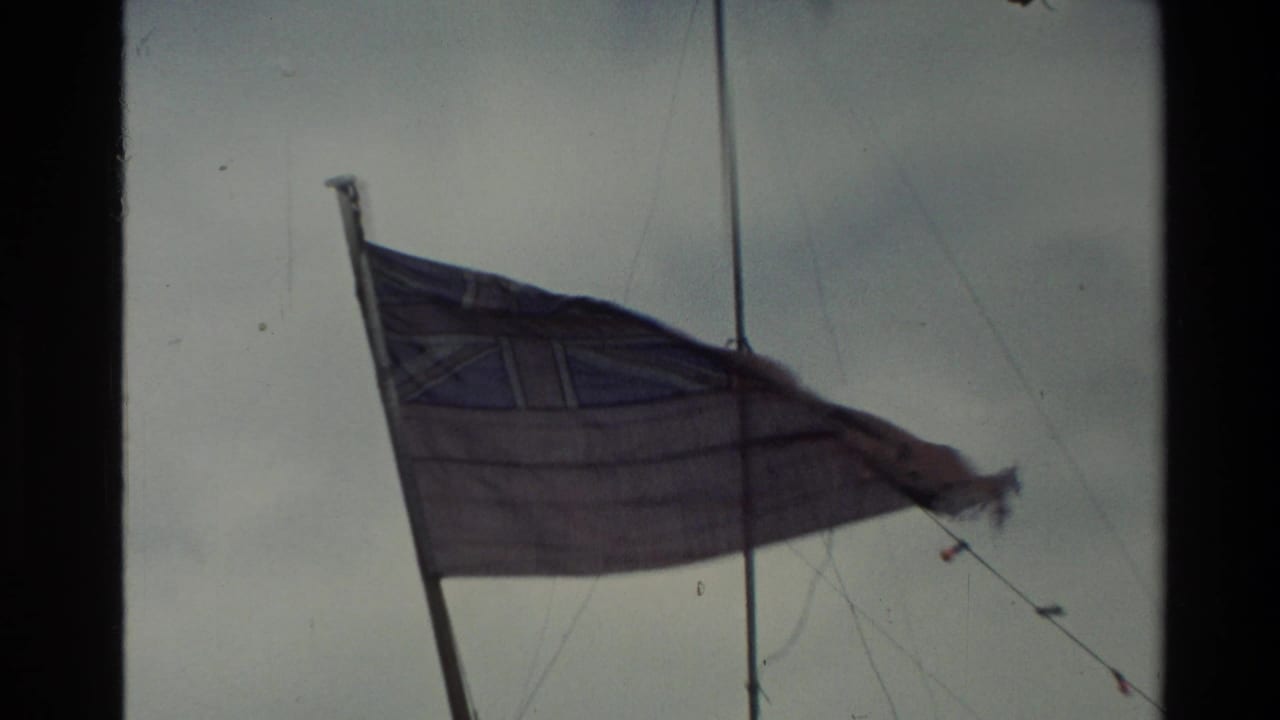 Faded flag, likely on a ferry in New Zealand, blowing in the wind against a pale cloudy sky in 1982 home-movie footage.