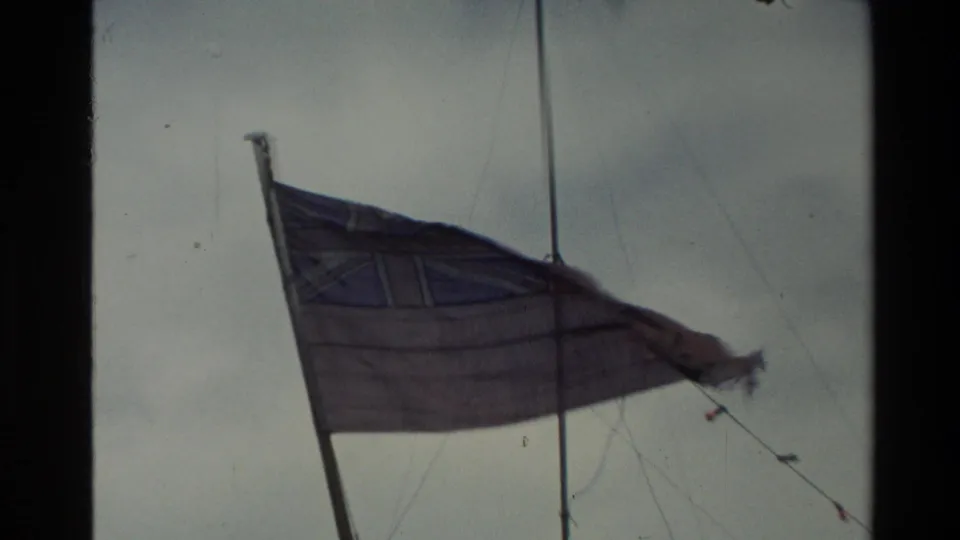 Faded flag, likely on a ferry in New Zealand, blowing in the wind against a pale cloudy sky in 1982 home-movie footage.