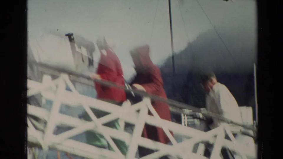 Passengers in raincoats appear to board a white ferry by gangway, with hillside and cloudy sky behind them in New Zealand footage.