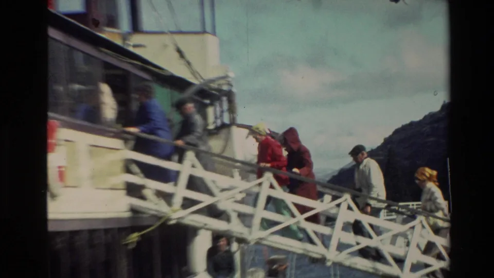 Several passengers board or stand near a white ferry gangway, with water, hillside, and cloudy sky visible in 1982 New Zealand footage.
