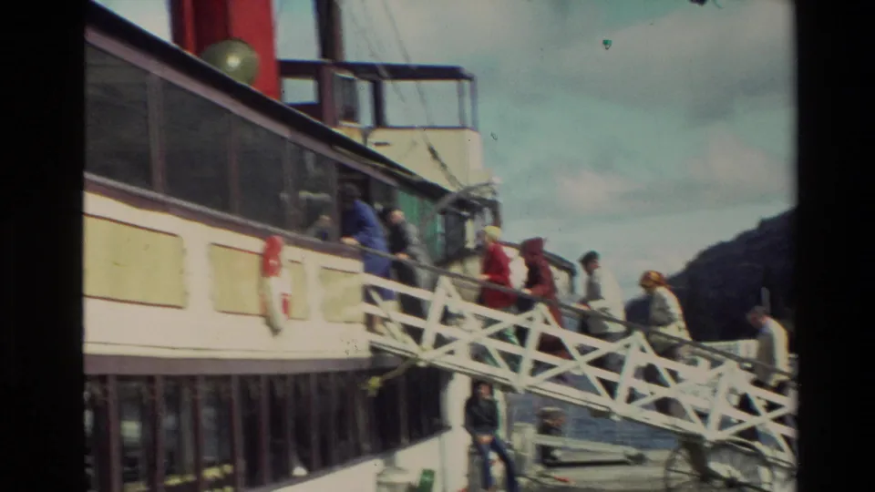 A wider view shows a white ferry, passengers on the gangway and deck, water, and a dark hillside in New Zealand home-movie footage.