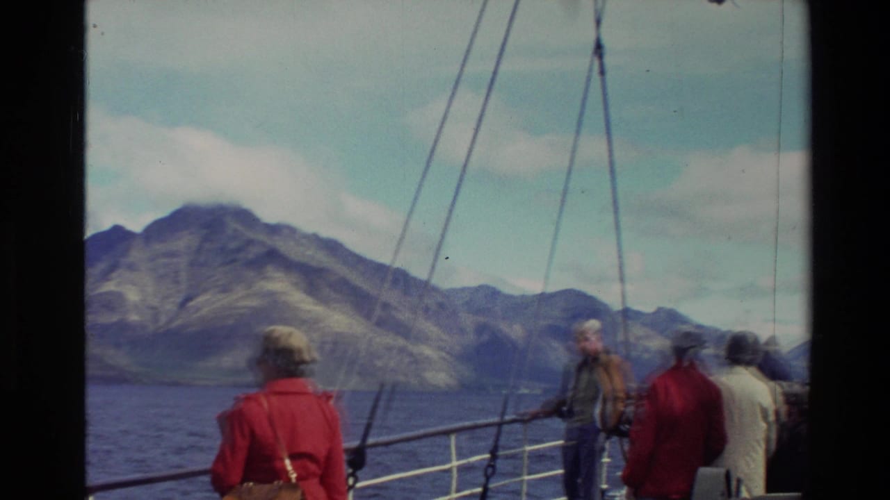 Tourists stand on a boat deck facing a mountain shoreline across open water.