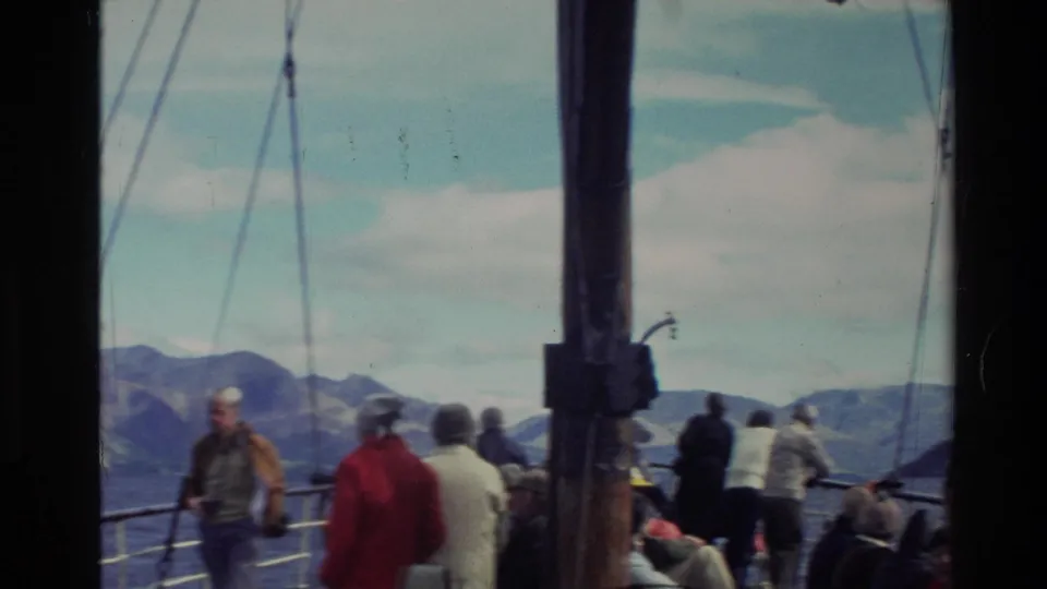 Tourists on a New Zealand sightseeing boat deck in 1982, with a dark mast, railings, calm water, and mountain slopes visible.