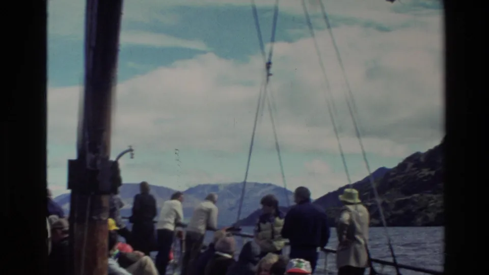 A visitor-shot 8mm view of tourists along a boat rail in New Zealand, with rigging overhead, water below, and mountains in the distance.