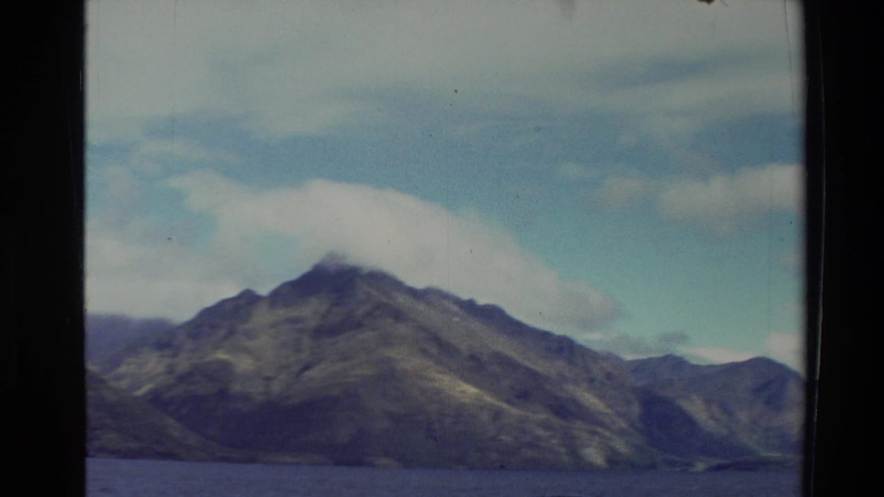 1982 New Zealand 8mm frame showing rugged mountains across lake water beneath broad white clouds and blue sky.