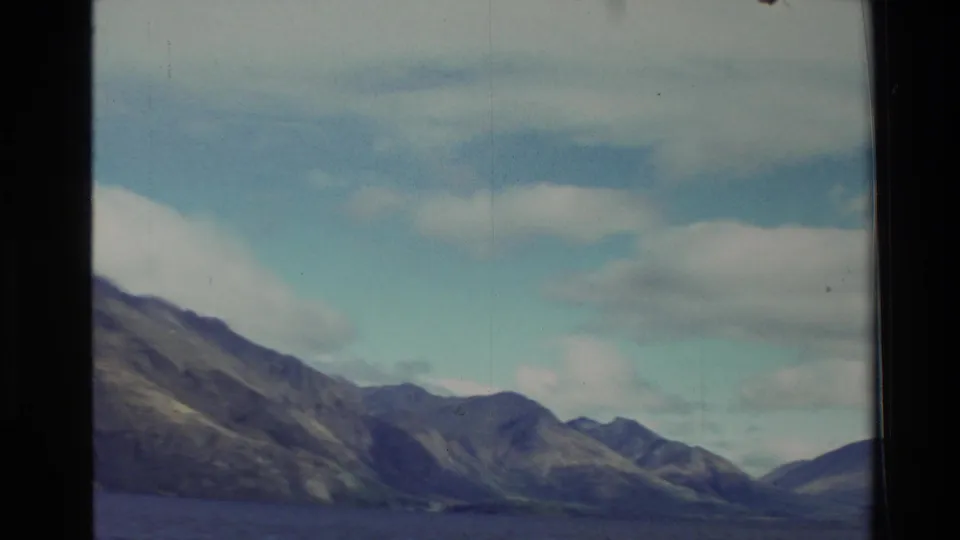 1982 New Zealand travel frame with lake water in the foreground and layered mountain slopes below white clouds.