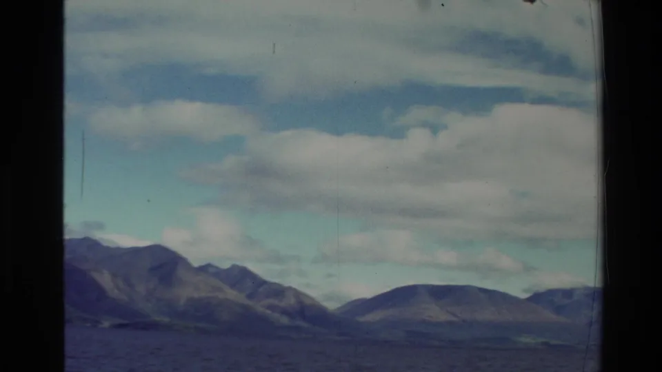 1982 New Zealand 8mm still showing a broad lake, distant mountain ridges, and large clouds from a moving boat viewpoint.
