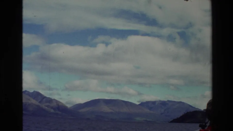 1982 New Zealand 8mm frame with distant mountains across the lake and a dark shoreline shape at the right edge.