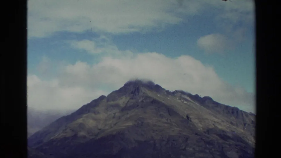1982 New Zealand 8mm frame showing a high green mountain peak partly covered by cloud under a blue sky.