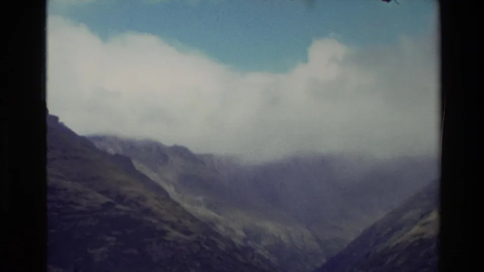 1982 New Zealand 8mm frame with layered mountain ridges and a valley partly hidden under broad white cloud.