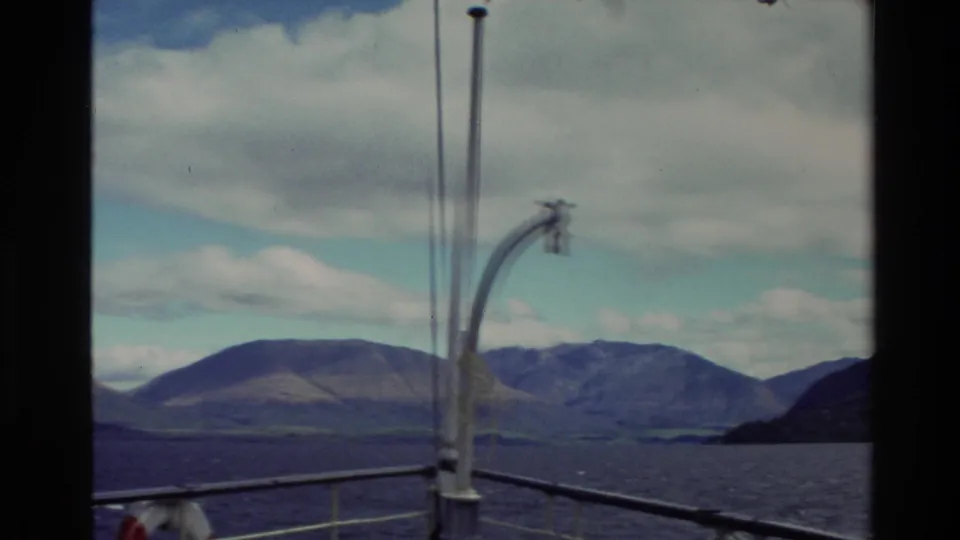 Deck-level view from a moving boat in New Zealand, showing railings, a curved lamp, open water, and mountain slopes.