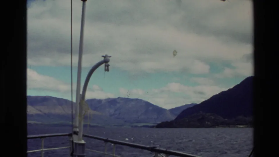 Handheld 8mm travel frame from a boat deck, with New Zealand mountains, shoreline, water, railings, and a curved deck lamp.