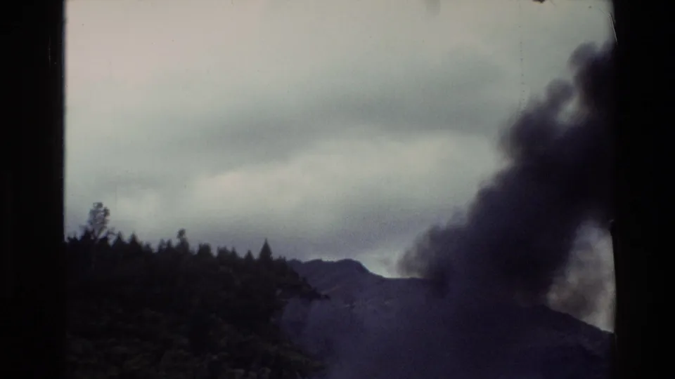 1982 New Zealand 8mm frame with smoke crossing a mountainous shoreline under an overcast sky.