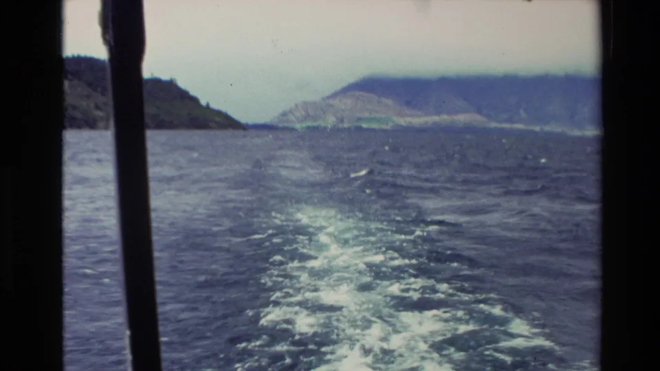 1982 New Zealand 8mm frame looking back over a foamy boat wake toward blue water and mountain shoreline.