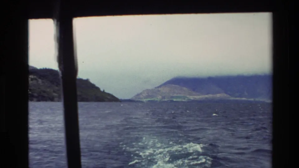 1982 New Zealand 8mm frame showing a boat's wake through deck posts, with mountainous shoreline beyond.