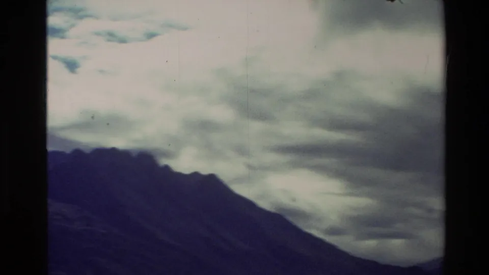 A dark New Zealand mountain ridge sits below layered overcast clouds in a soft, faded 8mm travel view.