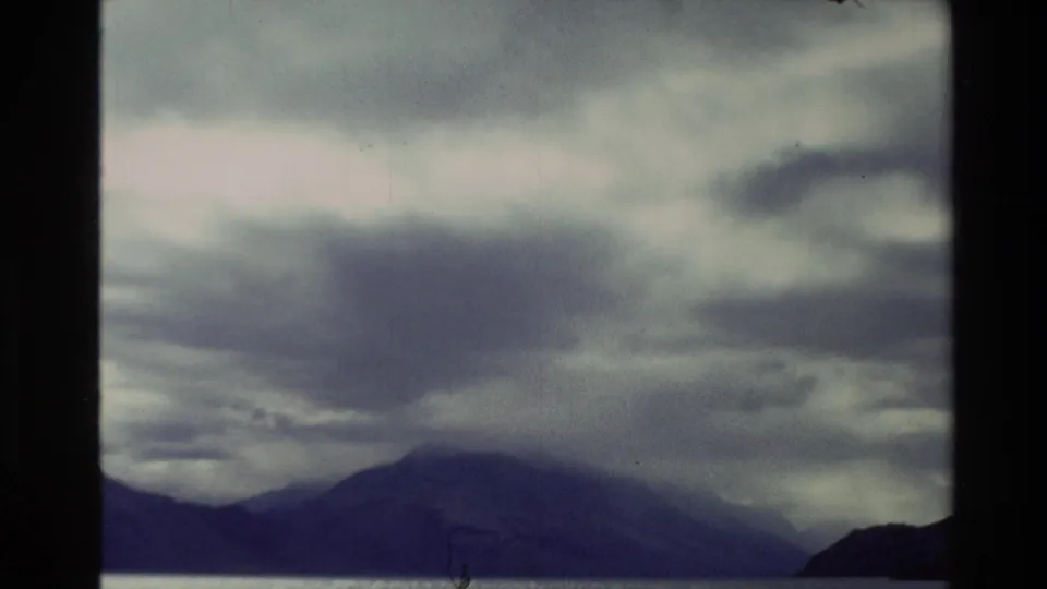 1982 New Zealand home-movie frame with a dark hill or mountain shape below heavy overcast clouds and muted 8mm color.