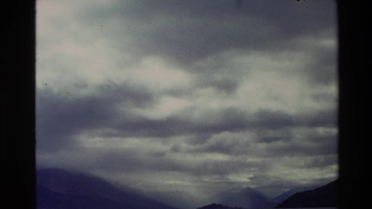 1982 New Zealand 8mm frame showing heavy cloud over dark mountain ridges and a subdued valley horizon.