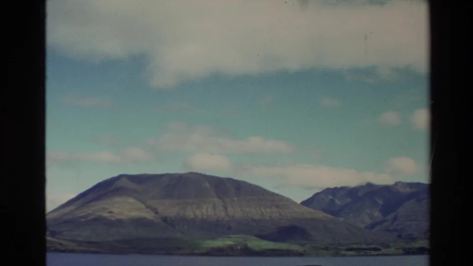 1982 New Zealand 8mm frame with rounded mountain forms, distant water, shoreline, and scattered clouds in a clear sky.