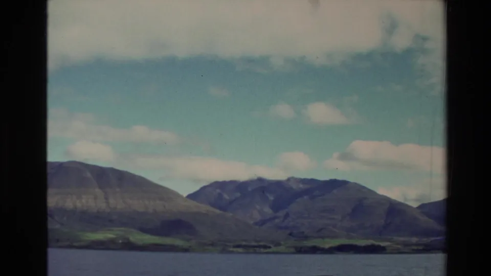 1982 New Zealand 8mm frame showing distant mountains, a low shoreline, and water beneath a partly cloudy blue sky.