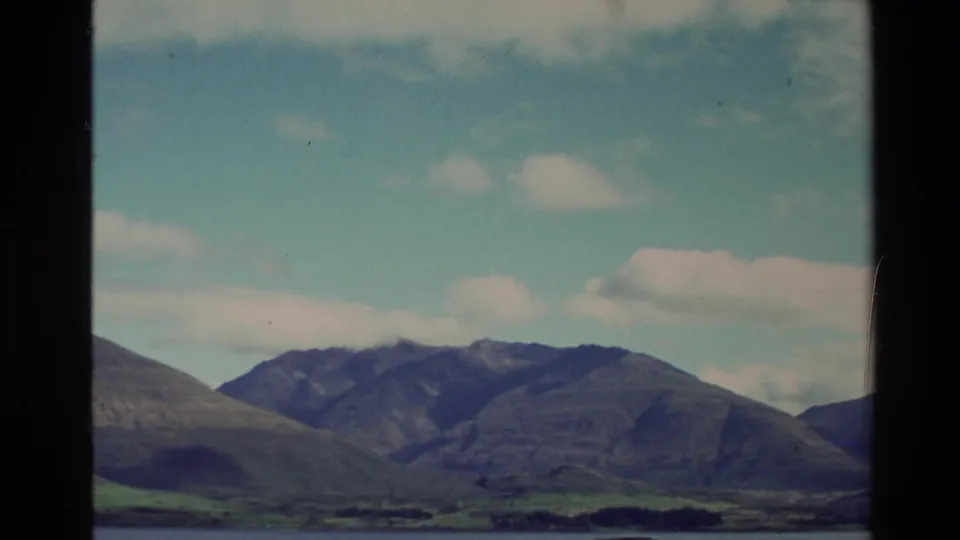 1982 New Zealand 8mm frame with a mountain skyline, grassy lower slopes, and small clouds across a bright blue sky.