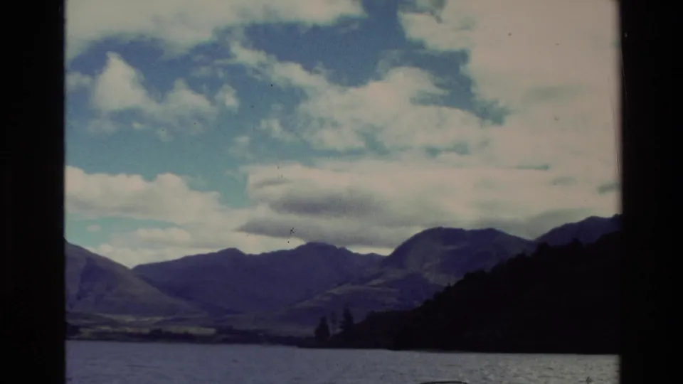 1982 New Zealand 8mm frame showing a mountain valley, lake surface, cloudy sky, and dark treeline.
