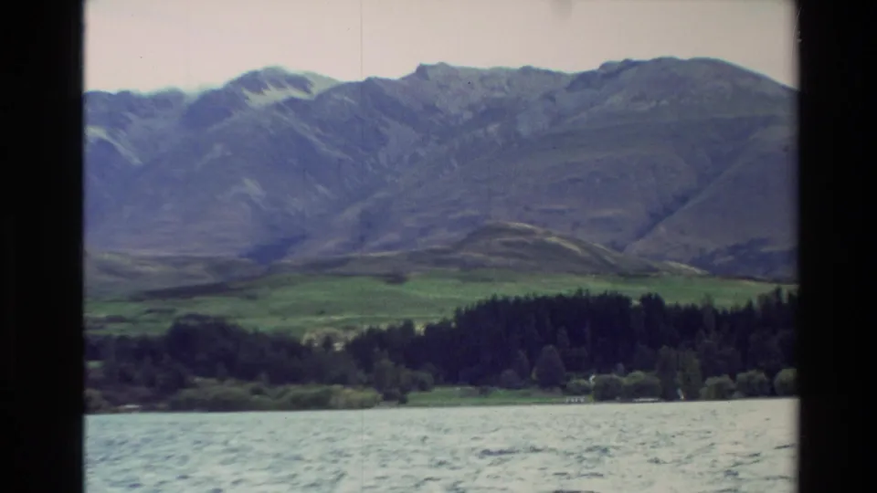 1982 New Zealand 8mm frame showing rippled lake water, a dark forest band, green valley, and mountains in the distance.
