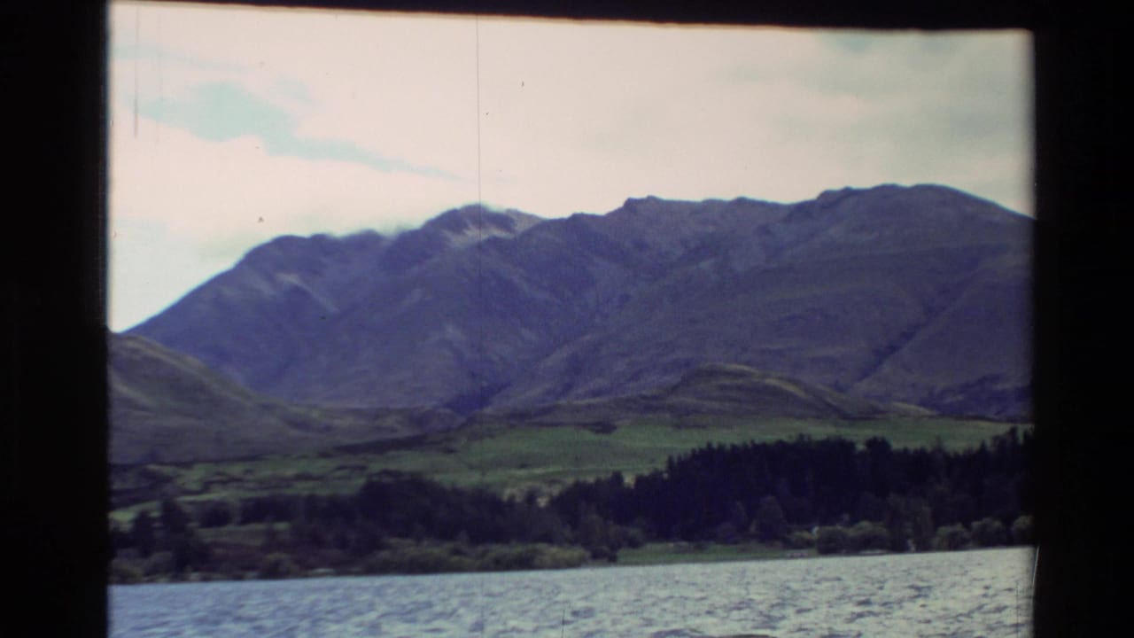 1982 New Zealand 8mm frame showing rippled water, wooded shoreline, green hills, and a broad mountain ridge under pale cloud.