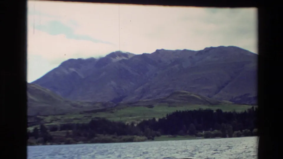1982 New Zealand 8mm frame showing rippled water, wooded shoreline, green hills, and a broad mountain ridge under pale cloud.