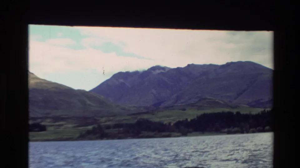 1982 New Zealand 8mm frame with water in the foreground, a wooded shore, green slopes, and blue-gray mountains in soft focus.