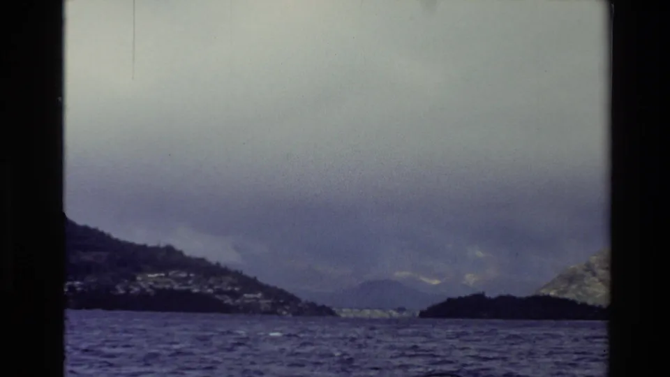 1982 New Zealand 8mm frame looking across darker water toward a distant shoreline, hills, and low clouds over mountain forms.