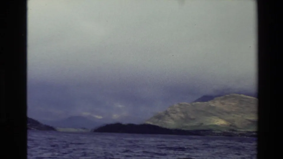 1982 New Zealand 8mm frame showing open water, a dark island or peninsula, distant hills, and a sunlit slope beneath layered cloud.