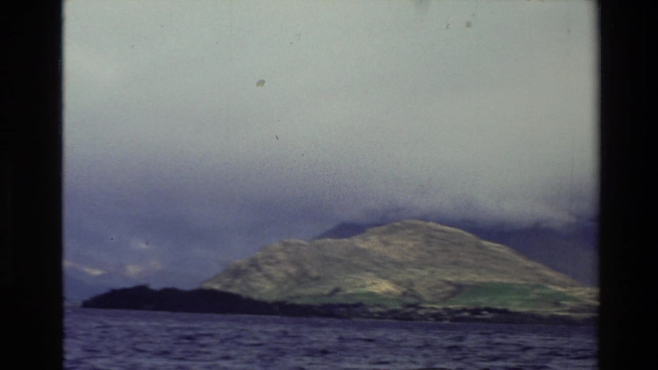 1982 New Zealand 8mm frame showing a broad lake and distant green hill partly covered by heavy cloud.