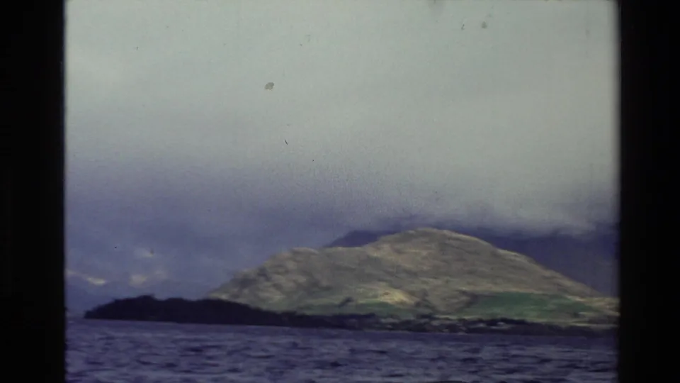 1982 New Zealand 8mm frame showing a broad lake and distant green hill partly covered by heavy cloud.
