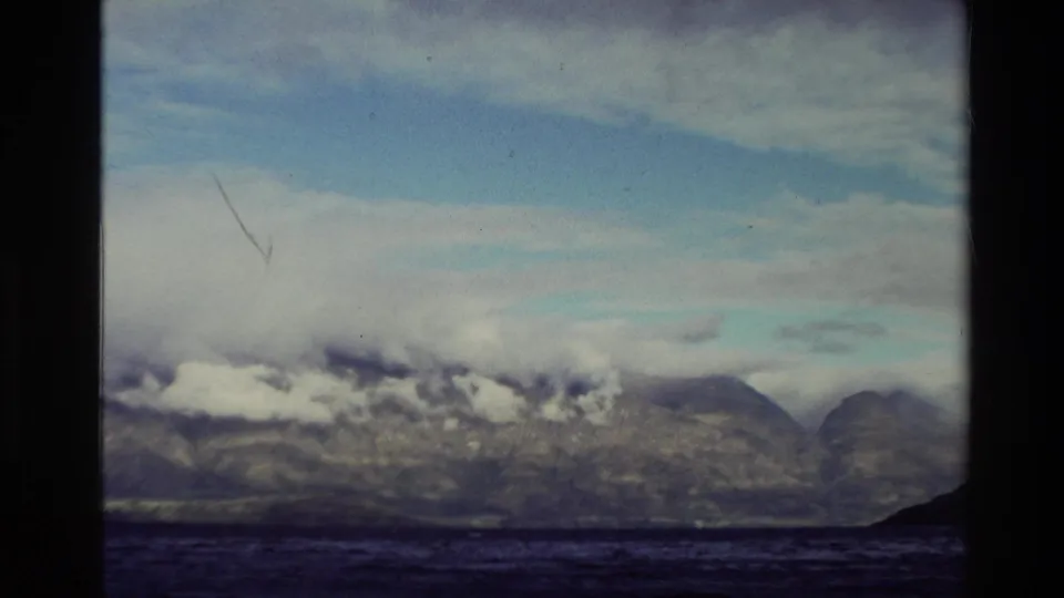 1982 New Zealand 8mm view of rocky mountains across a wide lake, with clouds hanging along the slopes.