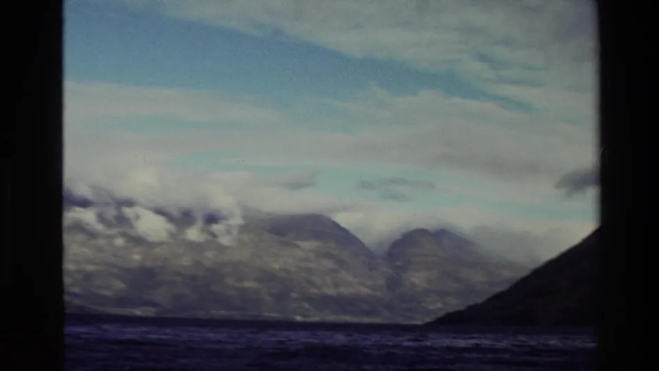 1982 New Zealand 8mm frame of distant cloud-covered mountains across water, bordered by a dark foreground shoreline.