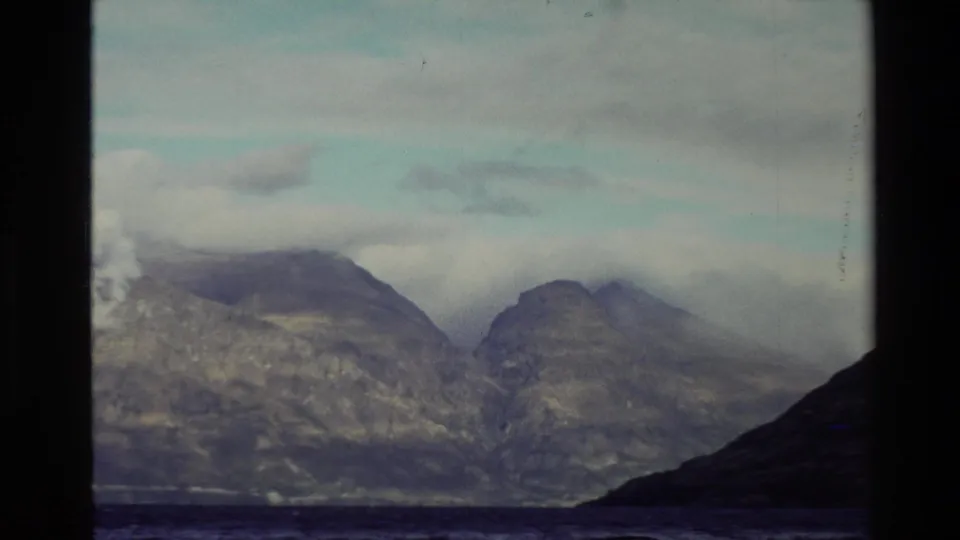 1982 New Zealand 8mm frame showing a lake, distant mountains and low cloud, with a dark shoreline rising at the right edge.