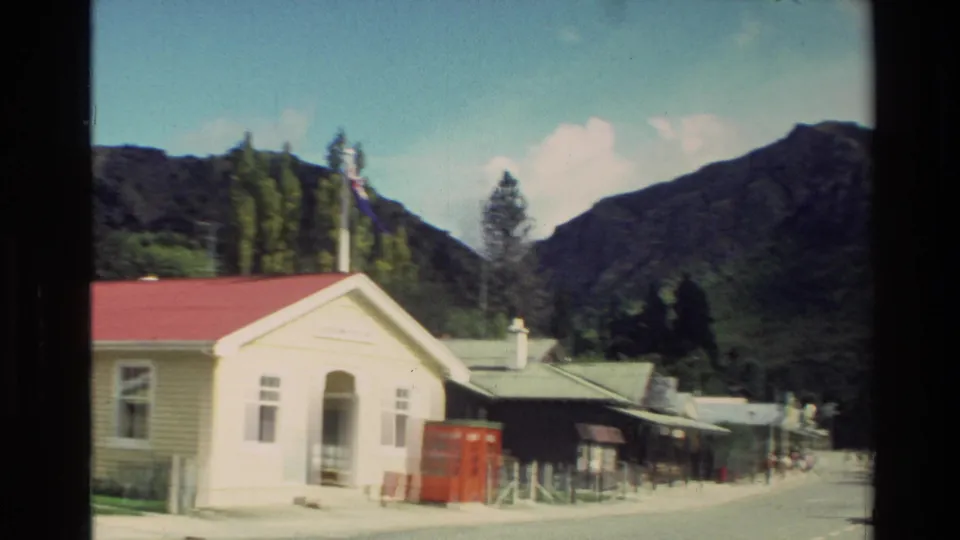 1982 New Zealand village street with a white red-roofed building, flagpole, and steep mountain backdrop.