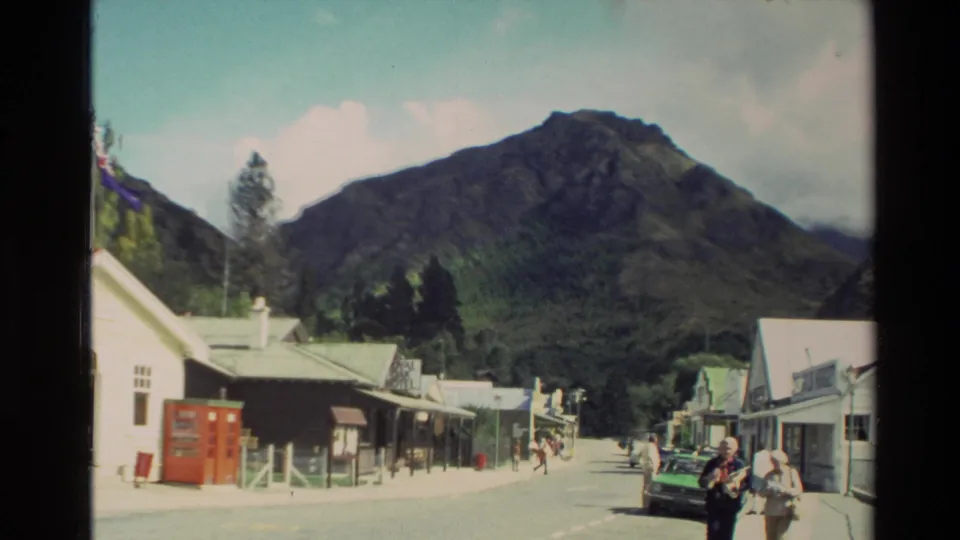 1982 New Zealand home-movie view of a small village street with pedestrians, shopfronts, a parked car, and mountains beyond.
