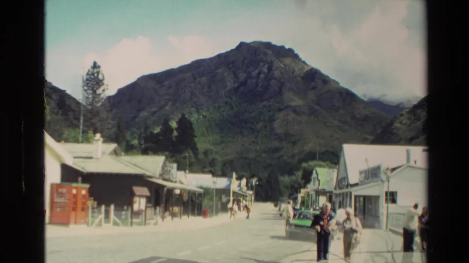 1982 New Zealand village street framed by mountains, with pedestrians walking near low buildings and a parked car.
