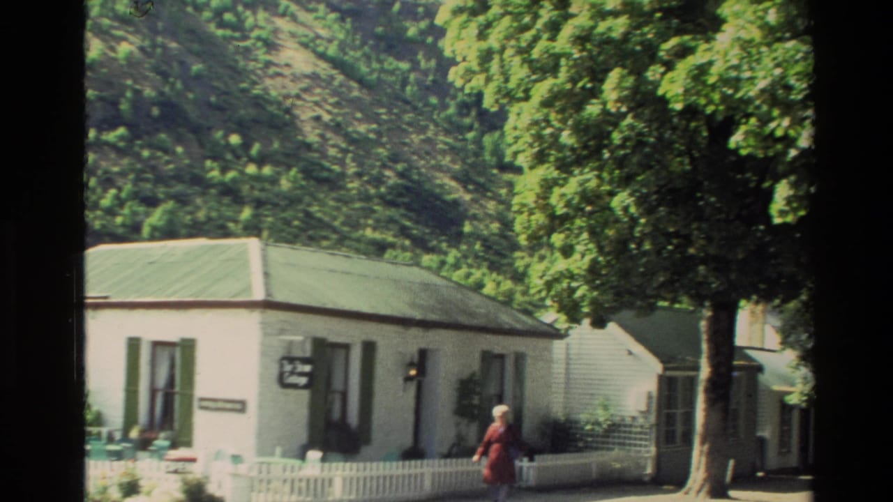 1982 New Zealand home-movie frame showing a woman in a red dress near white cottages, green shutters, and a tree-lined street.