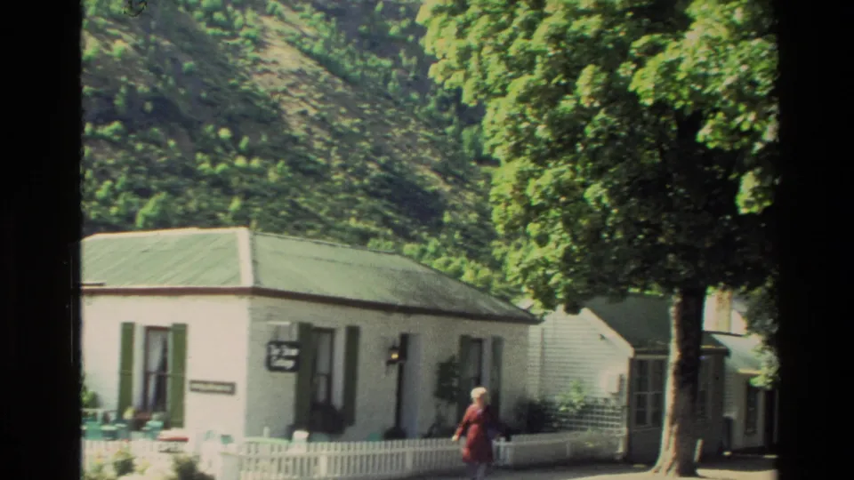 1982 New Zealand home-movie frame showing a woman in a red dress near white cottages, green shutters, and a tree-lined street.