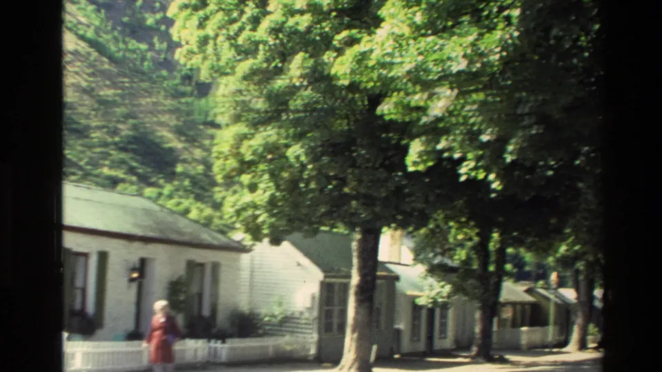 1982 New Zealand street view with a woman in red walking beside white cottages, fences, and dense summer trees.
