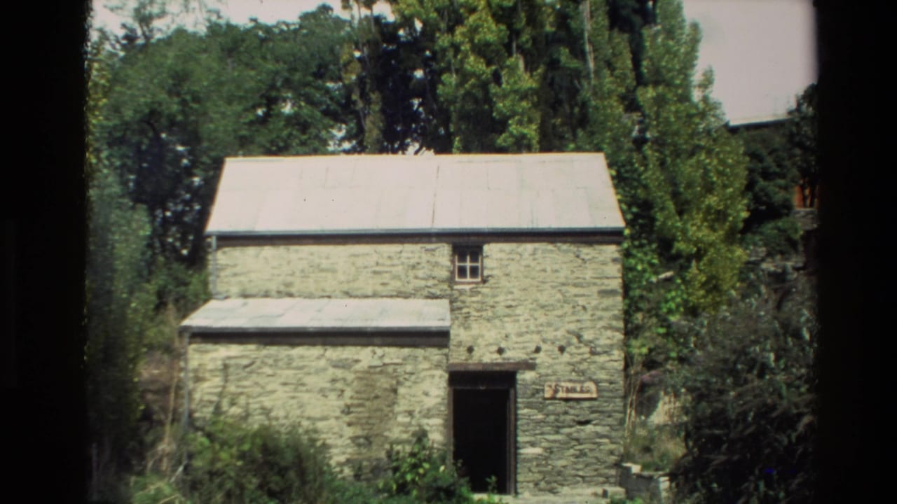 1982 New Zealand 8mm frame showing a weathered stone stable with a corrugated roof amid dense greenery.