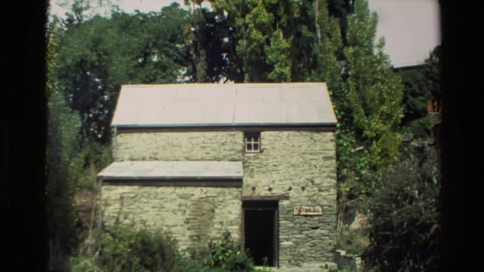 1982 New Zealand 8mm frame showing a weathered stone stable with a corrugated roof amid dense greenery.