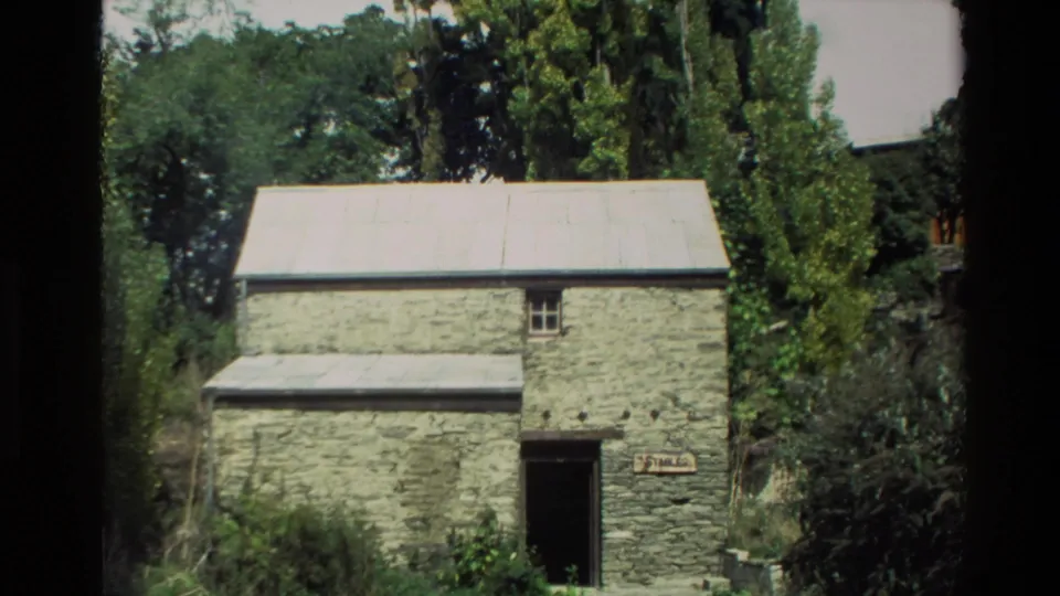 New Zealand home-movie frame of a rustic stone stable exterior, partly shaded by surrounding trees.