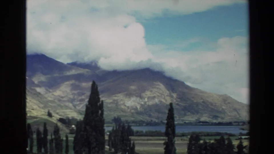 1982 New Zealand 8mm view of a mountain valley and lake with tall foreground trees and clouded peaks.