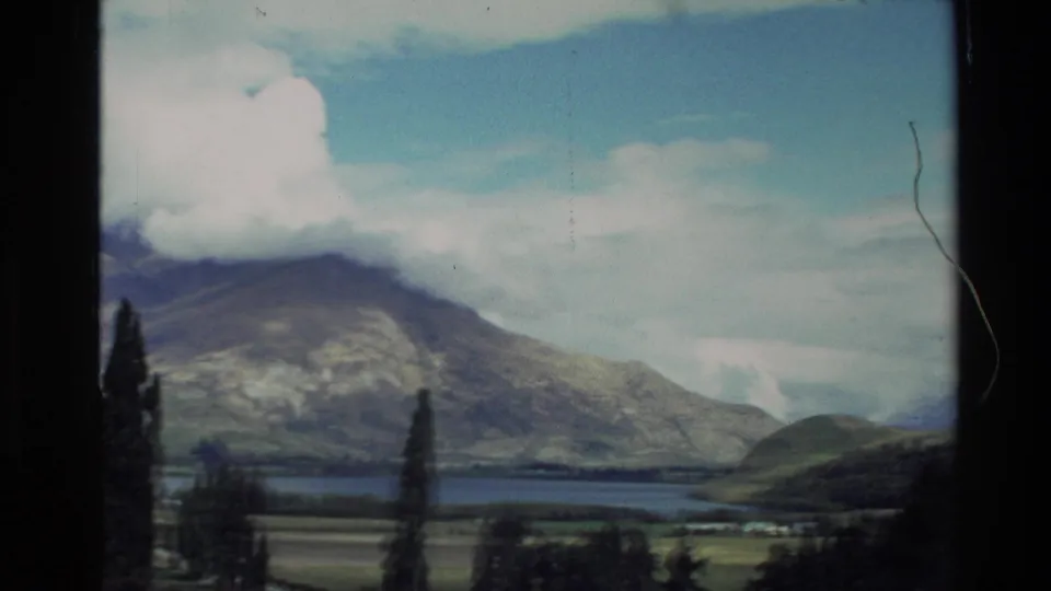 New Zealand travel-film frame showing a lake below broad mountain slopes, with cypress-like trees in front.